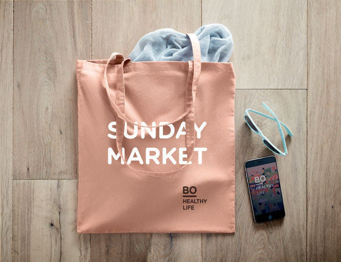 Pink tote bag with 'SUNDAY MARKET' text on a wooden floor, accompanied by a phone and headband.